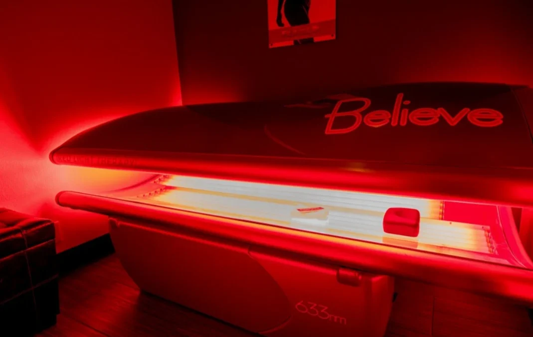 Red light therapy bed labeled Believe, open and glowing red, in a dimly lit room. Poster for LED Lipo on wall.
